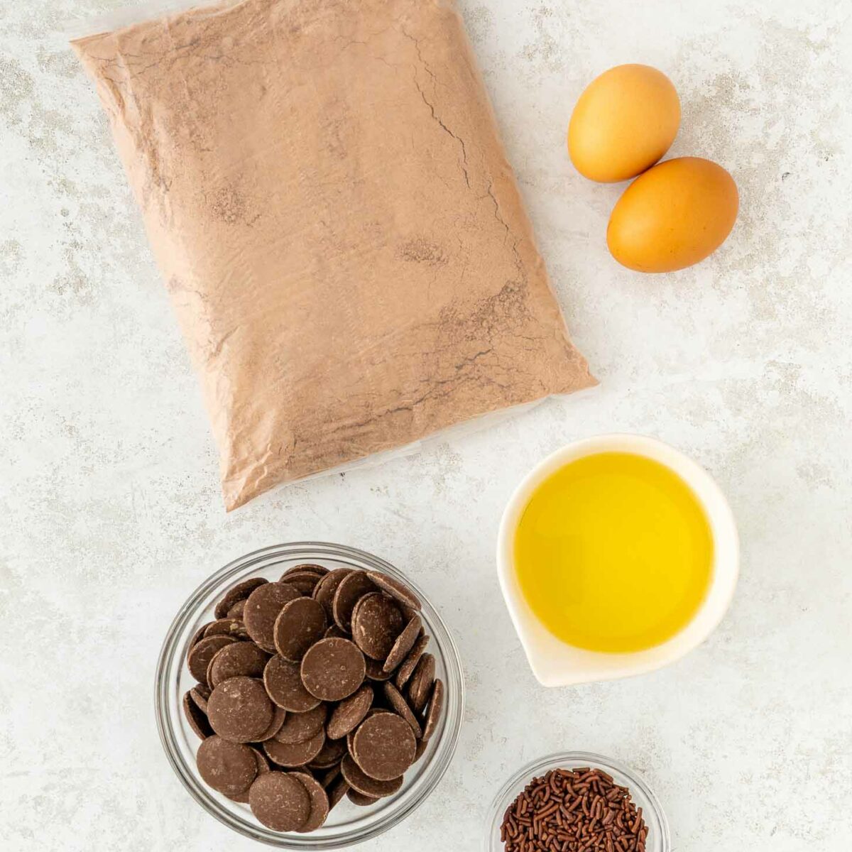 A top view of baking ingredients on a white surface: a bag of brownie mix, two eggs, a bowl of chocolate wafers, a bowl of chocolate sprinkles, and a small pitcher of yellow liquid, likely oil.