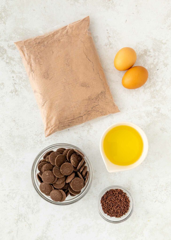 A top view of baking ingredients on a white surface: a bag of brownie mix, two eggs, a bowl of chocolate wafers, a bowl of chocolate sprinkles, and a small pitcher of yellow liquid, likely oil.