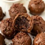 A close-up of brownie truffles covered in chocolate sprinkles, with one truffle showing a bite taken out, revealing a rich, fudgy center. A bowl of chocolate sprinkles is in the background.
