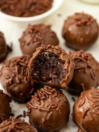 A close-up of brownie truffles covered in chocolate sprinkles, with one truffle showing a bite taken out, revealing a rich, fudgy center. A bowl of chocolate sprinkles is in the background.
