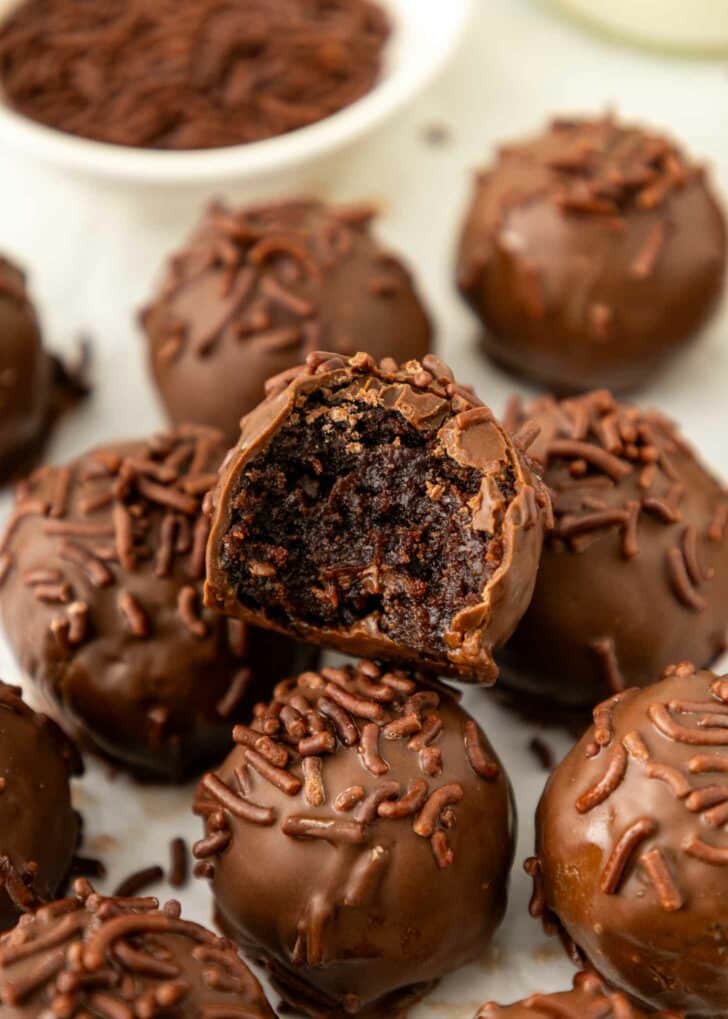 A close-up of brownie truffles covered in chocolate sprinkles, with one truffle showing a bite taken out, revealing a rich, fudgy center. A bowl of chocolate sprinkles is in the background.