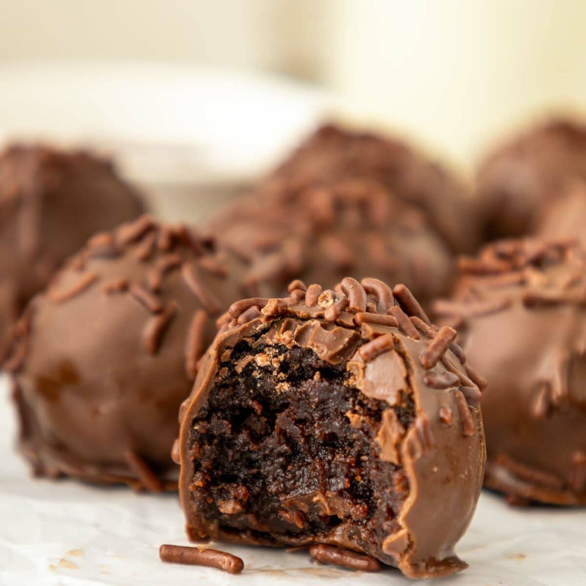Close-up of brownie truffles covered in chocolate sprinkles, with one truffle in front showing a bite taken out, revealing a rich, fudgy center. The background is slightly blurred.