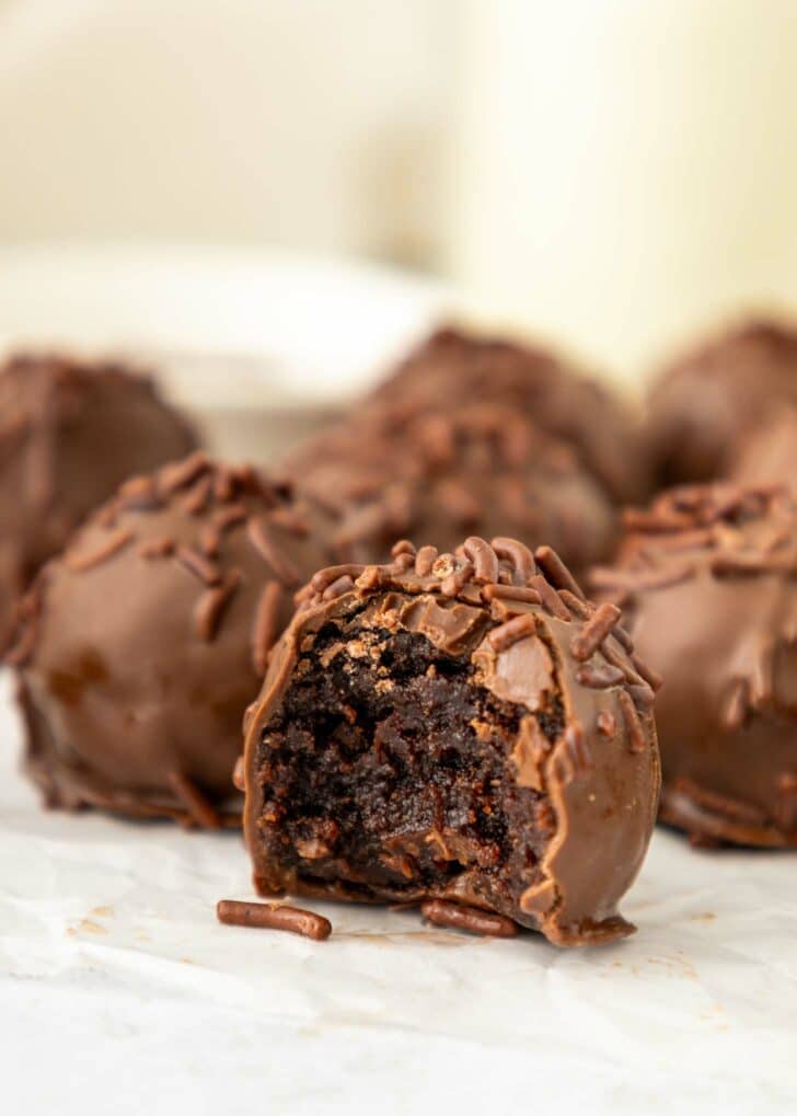 Close-up of brownie truffles covered in chocolate sprinkles, with one truffle in front showing a bite taken out, revealing a rich, fudgy center. The background is slightly blurred.