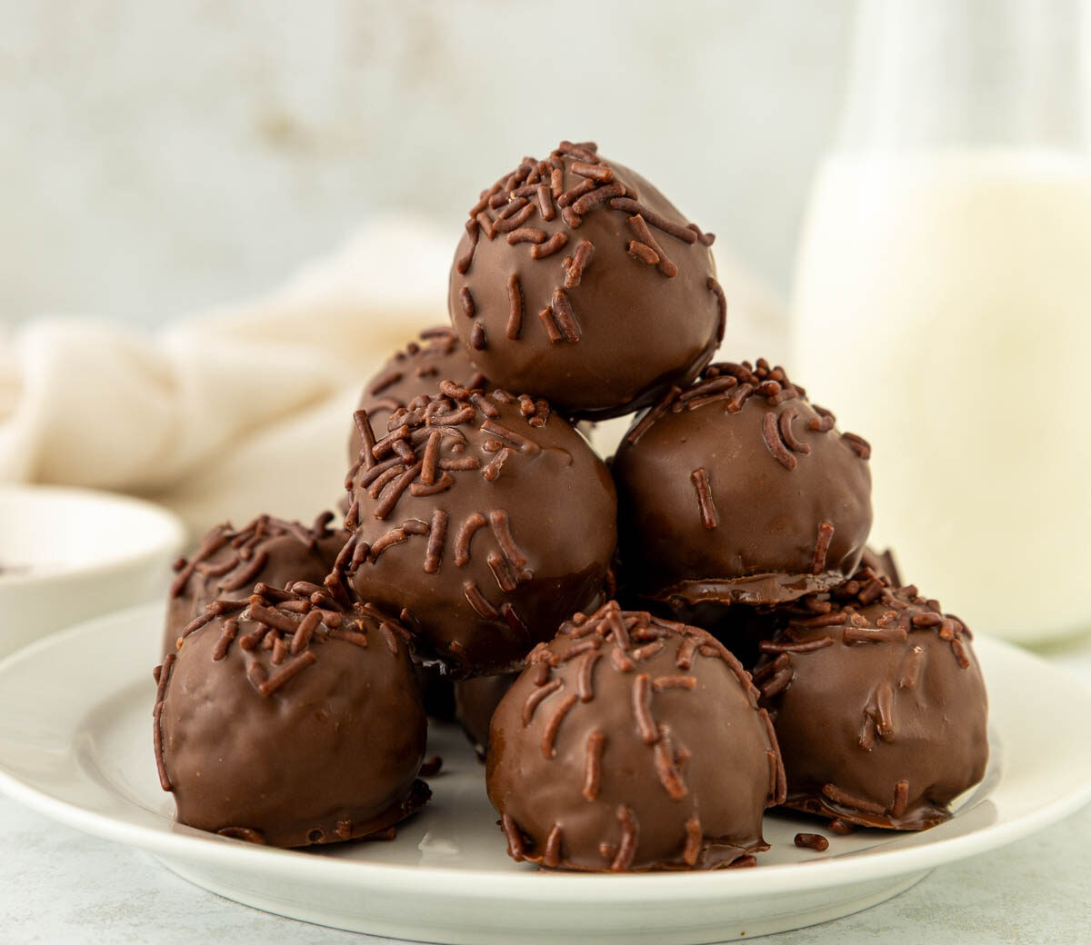 A plate stacked with round brownie truffles covered in chocolate sprinkles, with a milk bottle and a bowl in the soft-focus background.