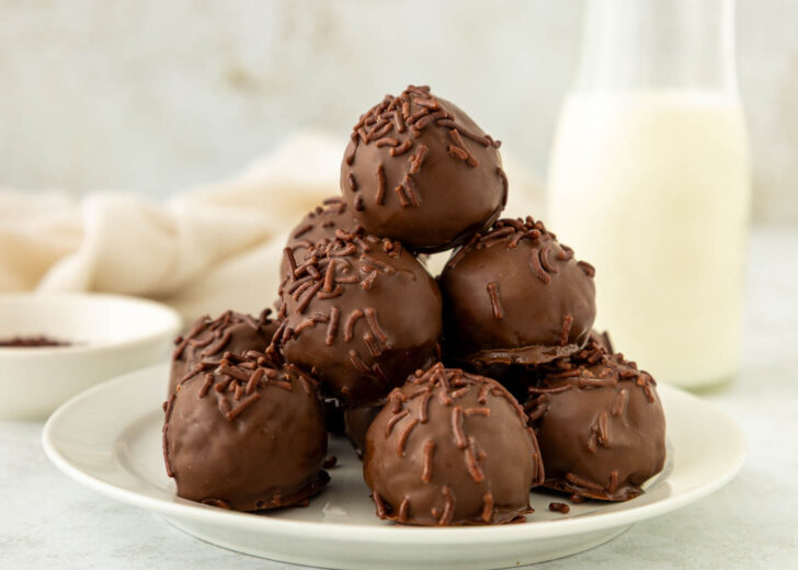 A plate stacked with round brownie truffles covered in chocolate sprinkles, with a milk bottle and a bowl in the soft-focus background.