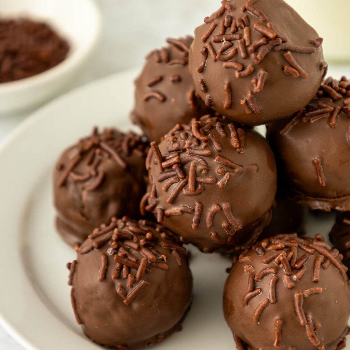 A close-up of a white plate stacked with round brownie truffles coated in chocolate and topped with chocolate sprinkles. A small bowl with more chocolate sprinkles is in the background.