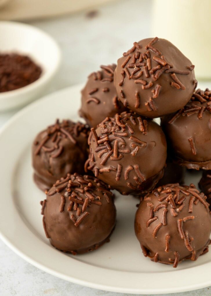 A close-up of a white plate stacked with round brownie truffles coated in chocolate and topped with chocolate sprinkles. A small bowl with more chocolate sprinkles is in the background.