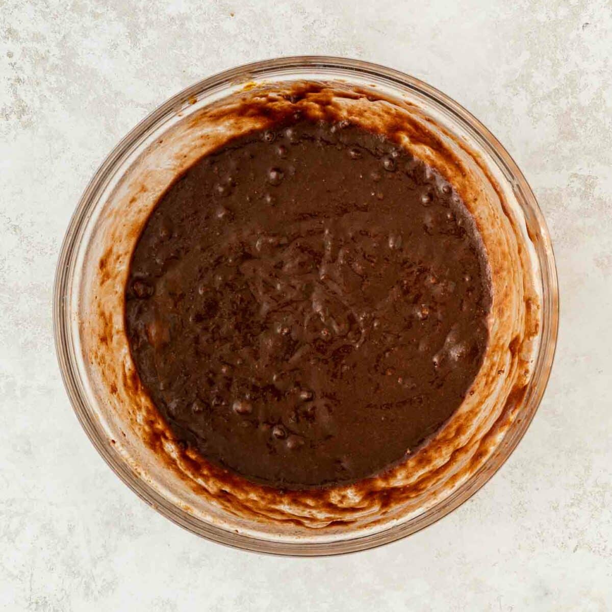 A glass bowl filled with thick, chocolate brownie batter sits on a light textured surface. The batter is dark brown and swirled, showing some unmixed streaks along the sides of the bowl.