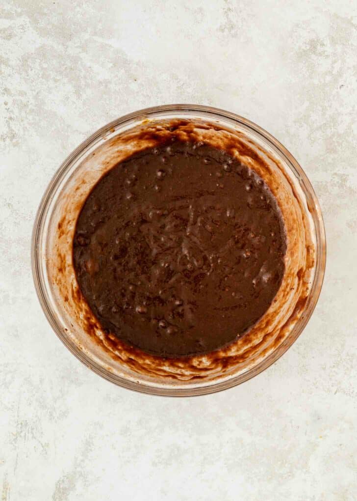 A glass bowl filled with thick, chocolate brownie batter sits on a light textured surface. The batter is dark brown and swirled, showing some unmixed streaks along the sides of the bowl.