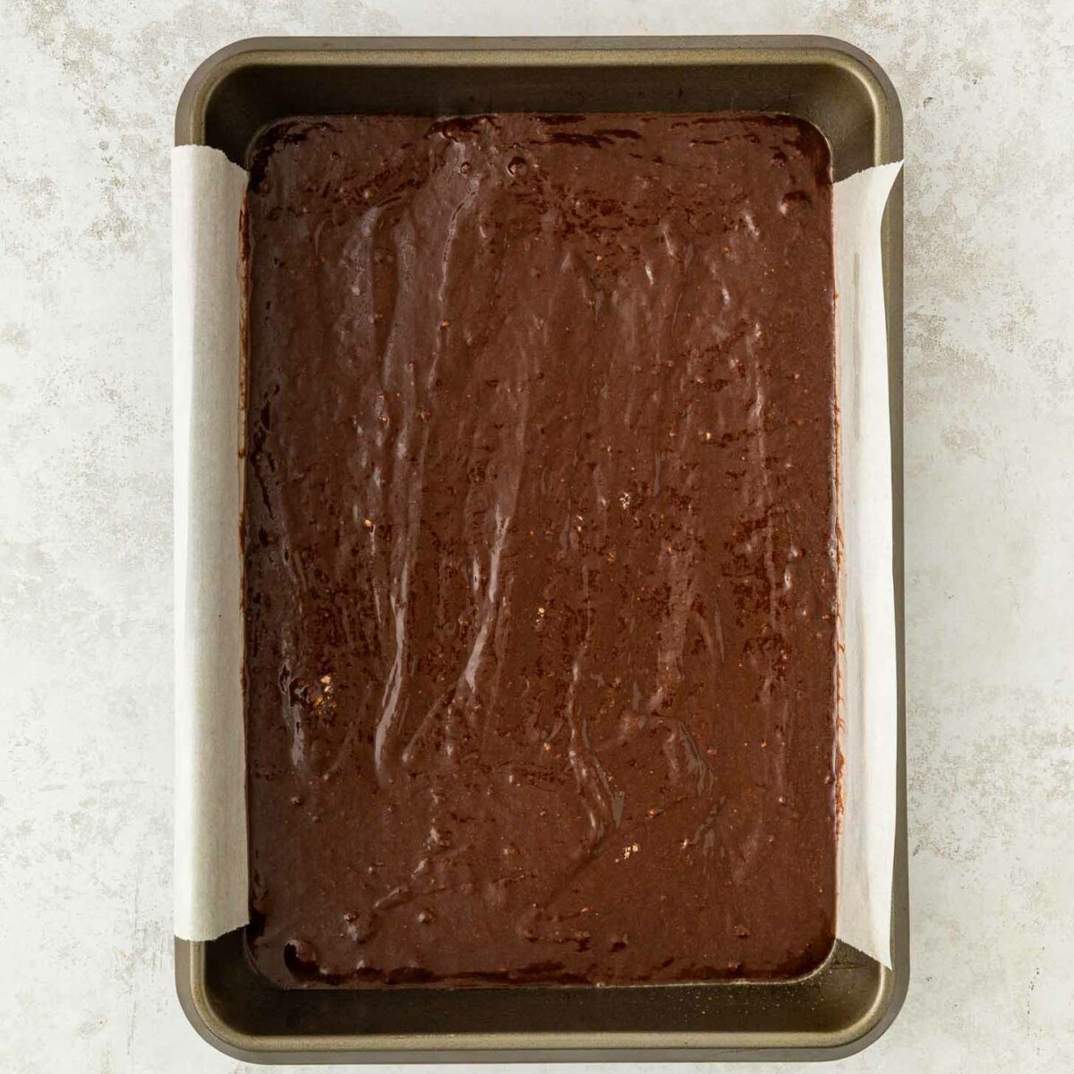 A rectangular metal baking pan lined with parchment paper, filled with unbaked chocolate brownie batter, sits on a light textured surface.