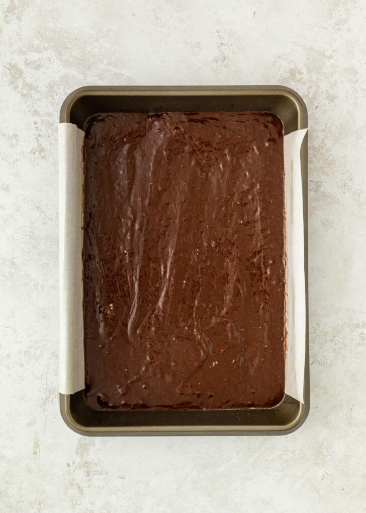 A rectangular metal baking pan lined with parchment paper, filled with unbaked chocolate brownie batter, sits on a light textured surface.