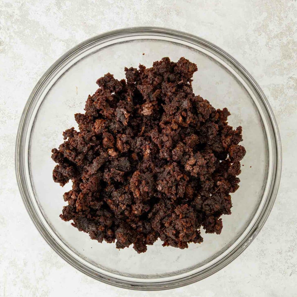 A clear glass bowl filled with crumbled chocolate brownies sits on a light, textured surface.
