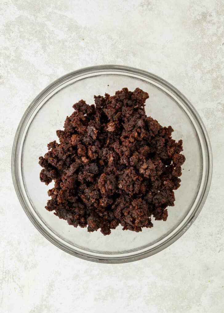 A clear glass bowl filled with crumbled chocolate brownies sits on a light, textured surface.