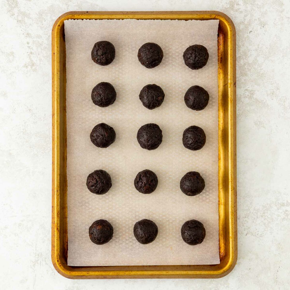 A baking tray lined with parchment paper holds fifteen evenly spaced balls of dark chocolate brownie truffles, arranged in a 3 by 5 grid, on a light surface.