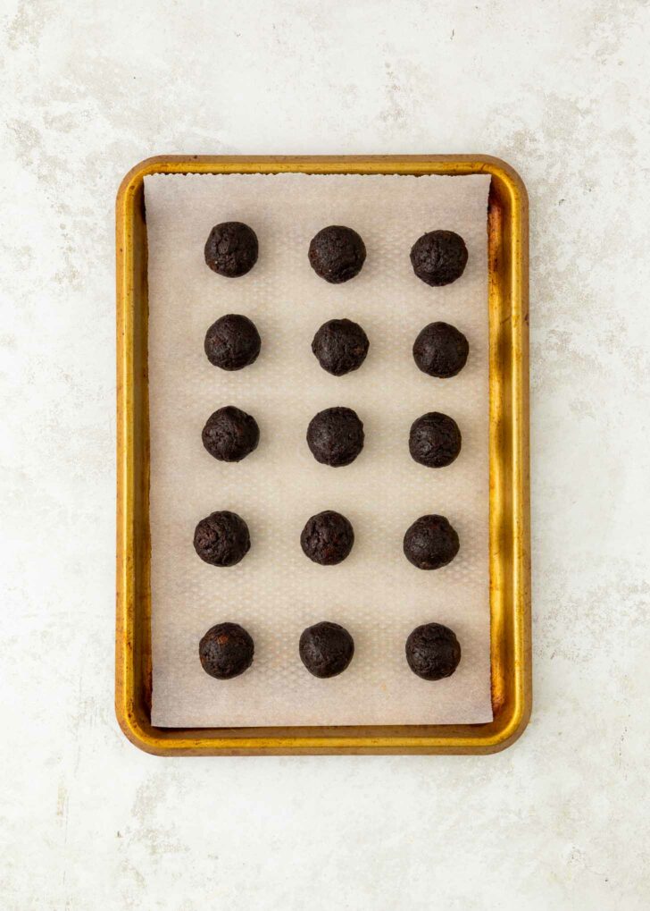 A baking tray lined with parchment paper holds fifteen evenly spaced balls of dark chocolate brownie truffles, arranged in a 3 by 5 grid, on a light surface.