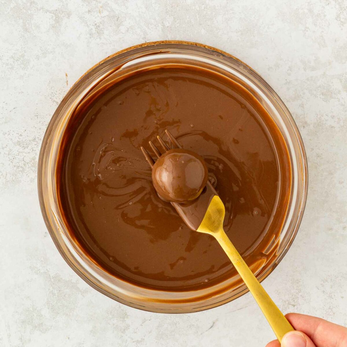A hand holding a gold fork dips a round treat into a bowl of melted chocolate, fully coating it. The bowl sits on a light, textured surface.
