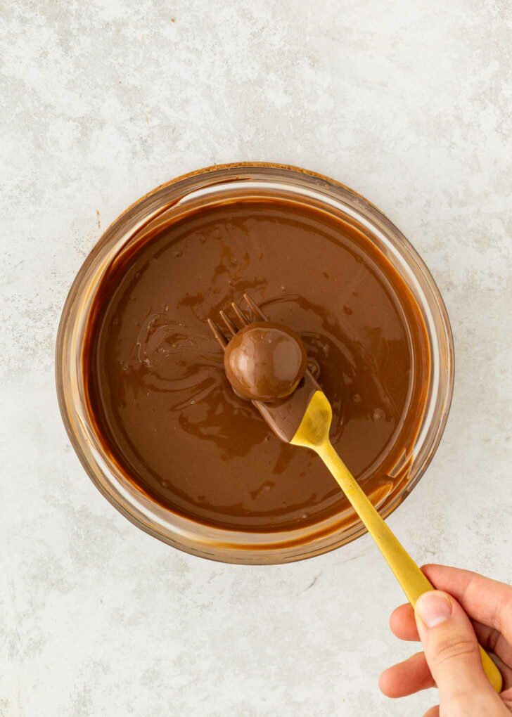 A hand holding a gold fork dips a round treat into a bowl of melted chocolate, fully coating it. The bowl sits on a light, textured surface.