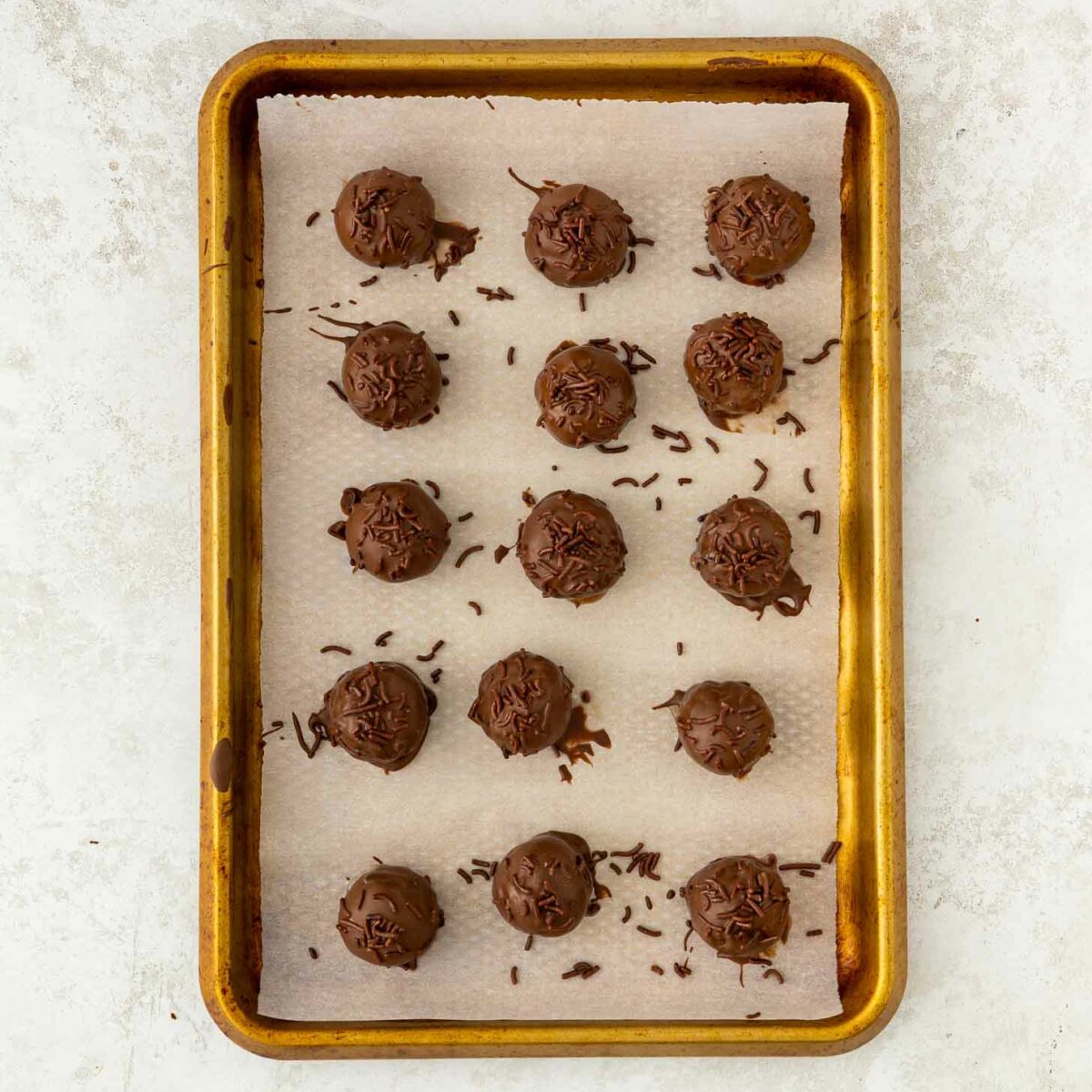 A baking sheet lined with parchment paper holds 15 round brownie truffles, each topped with chocolate shavings, arranged neatly in rows on a light-colored surface.