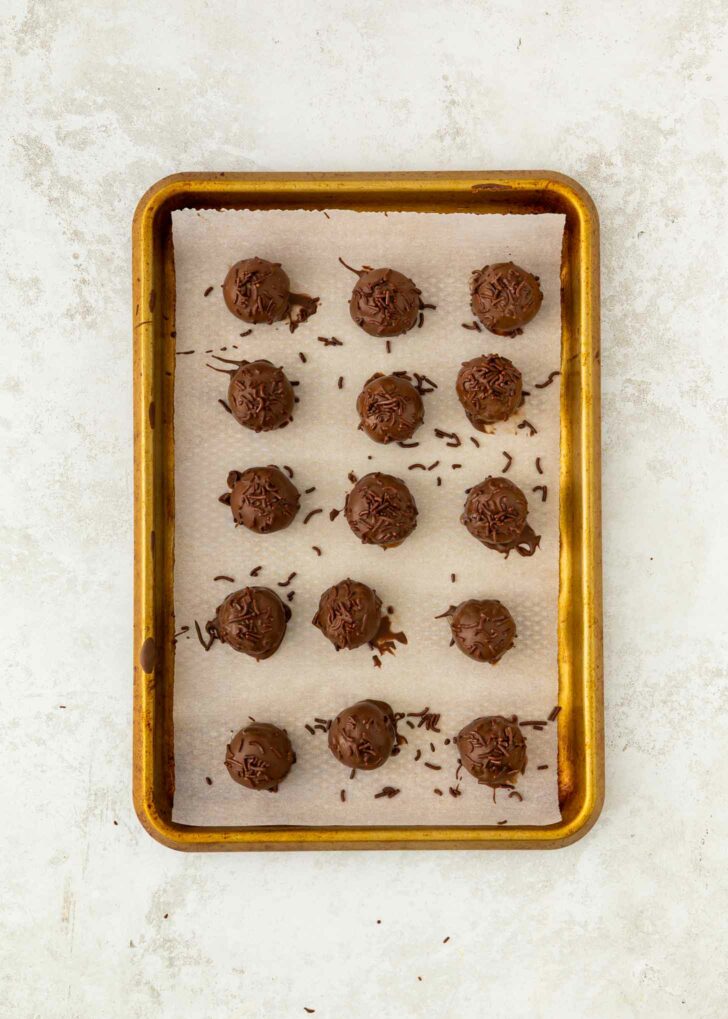 A baking sheet lined with parchment paper holds 15 round brownie truffles, each topped with chocolate shavings, arranged neatly in rows on a light-colored surface.