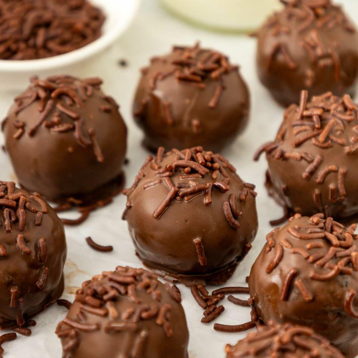 Brownie truffles coated in chocolate sprinkles are arranged on a white surface, with a bowl of extra sprinkles and a blurred glass of milk in the background.