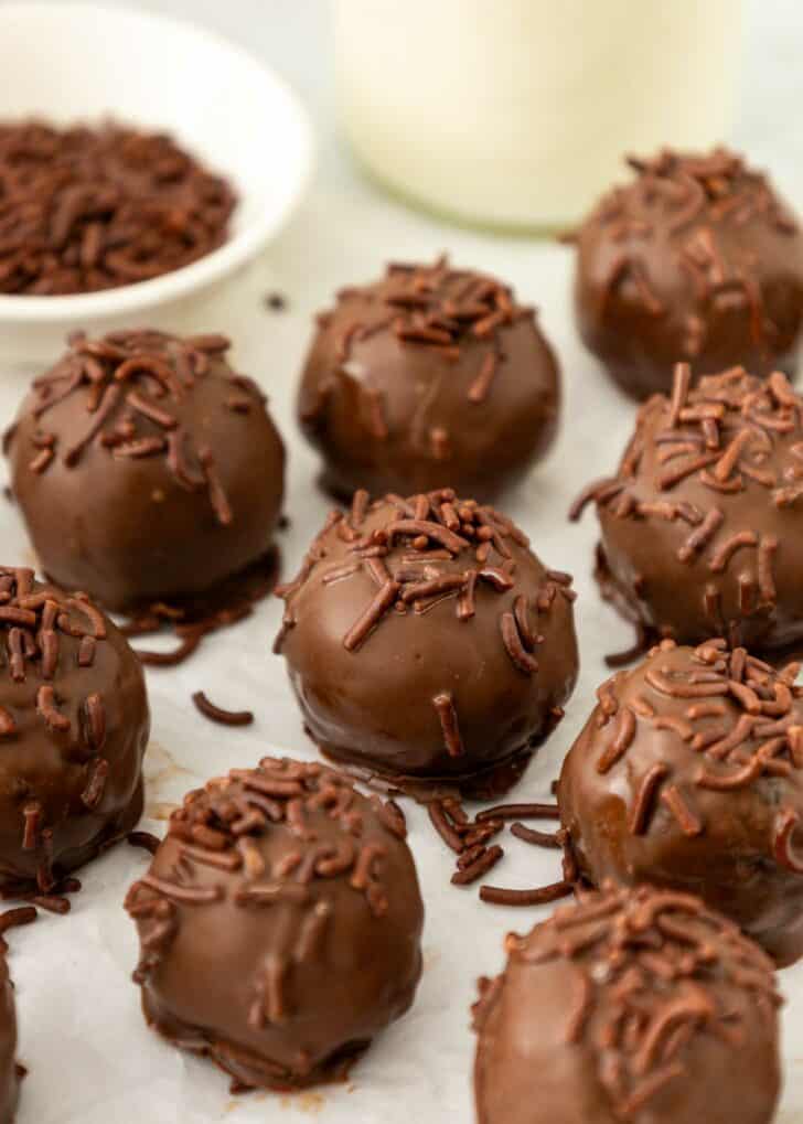 Brownie truffles coated in chocolate sprinkles are arranged on a white surface, with a bowl of extra sprinkles and a blurred glass of milk in the background.