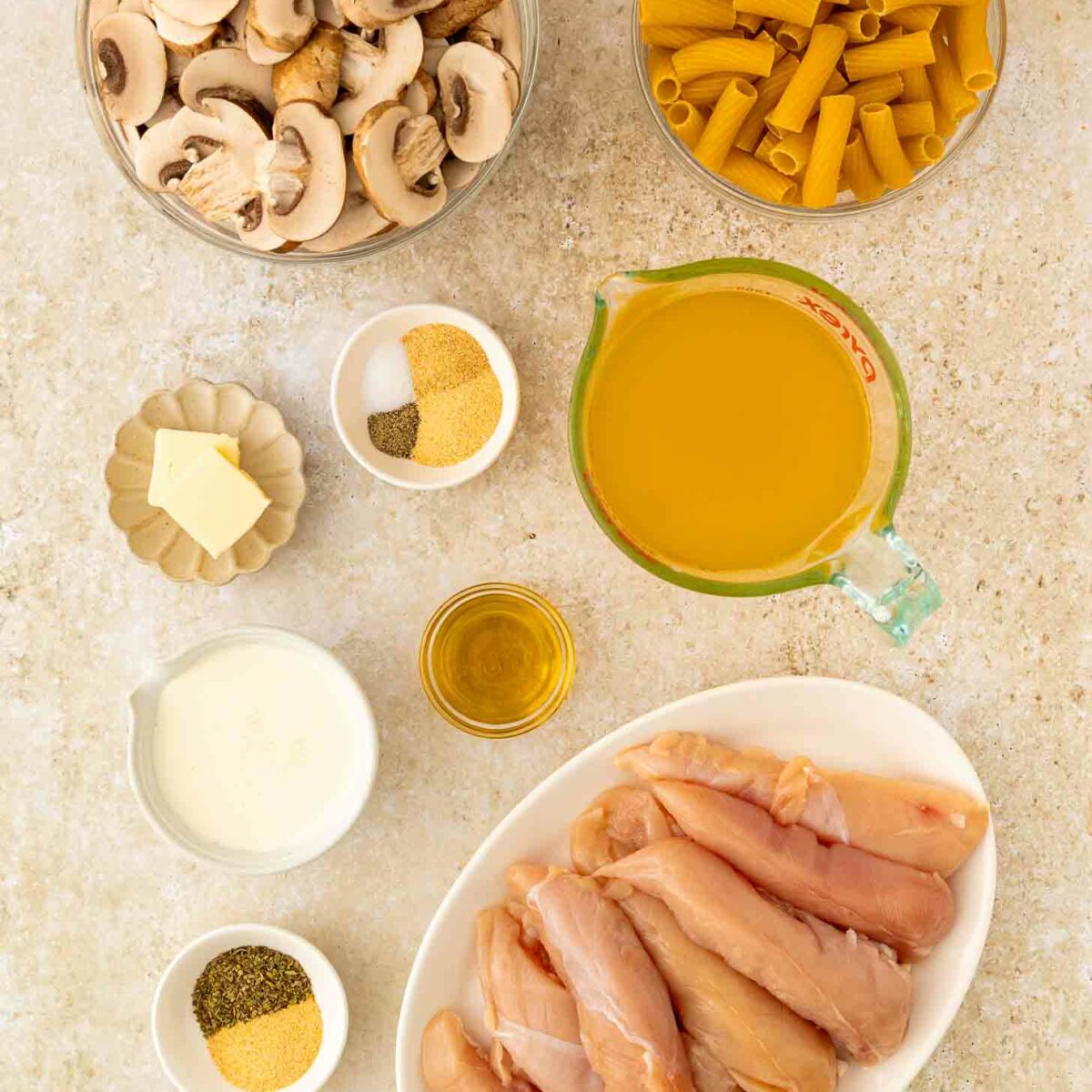 Top-down view of ingredients for a chicken and pasta dish, including raw chicken tenders, sliced mushrooms, uncooked pasta, chicken broth, butter, olive oil, cream, and various spices in small bowls.