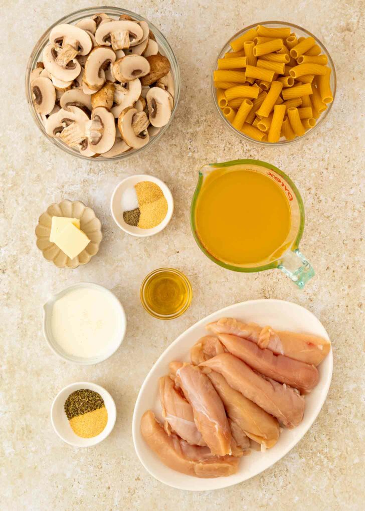Top-down view of ingredients for a chicken and pasta dish, including raw chicken tenders, sliced mushrooms, uncooked pasta, chicken broth, butter, olive oil, cream, and various spices in small bowls.