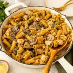 A skillet filled with rigatoni pasta, chicken pieces, mushrooms, and parsley, topped with grated cheese. A wooden spoon rests in the pan, with plates, utensils, and fresh parsley on the side.