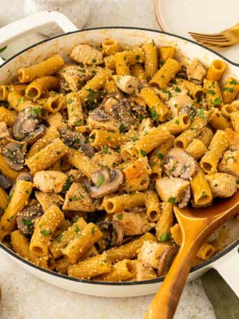 A skillet filled with rigatoni pasta, chicken pieces, mushrooms, and parsley, topped with grated cheese. A wooden spoon rests in the pan, with plates, utensils, and fresh parsley on the side.