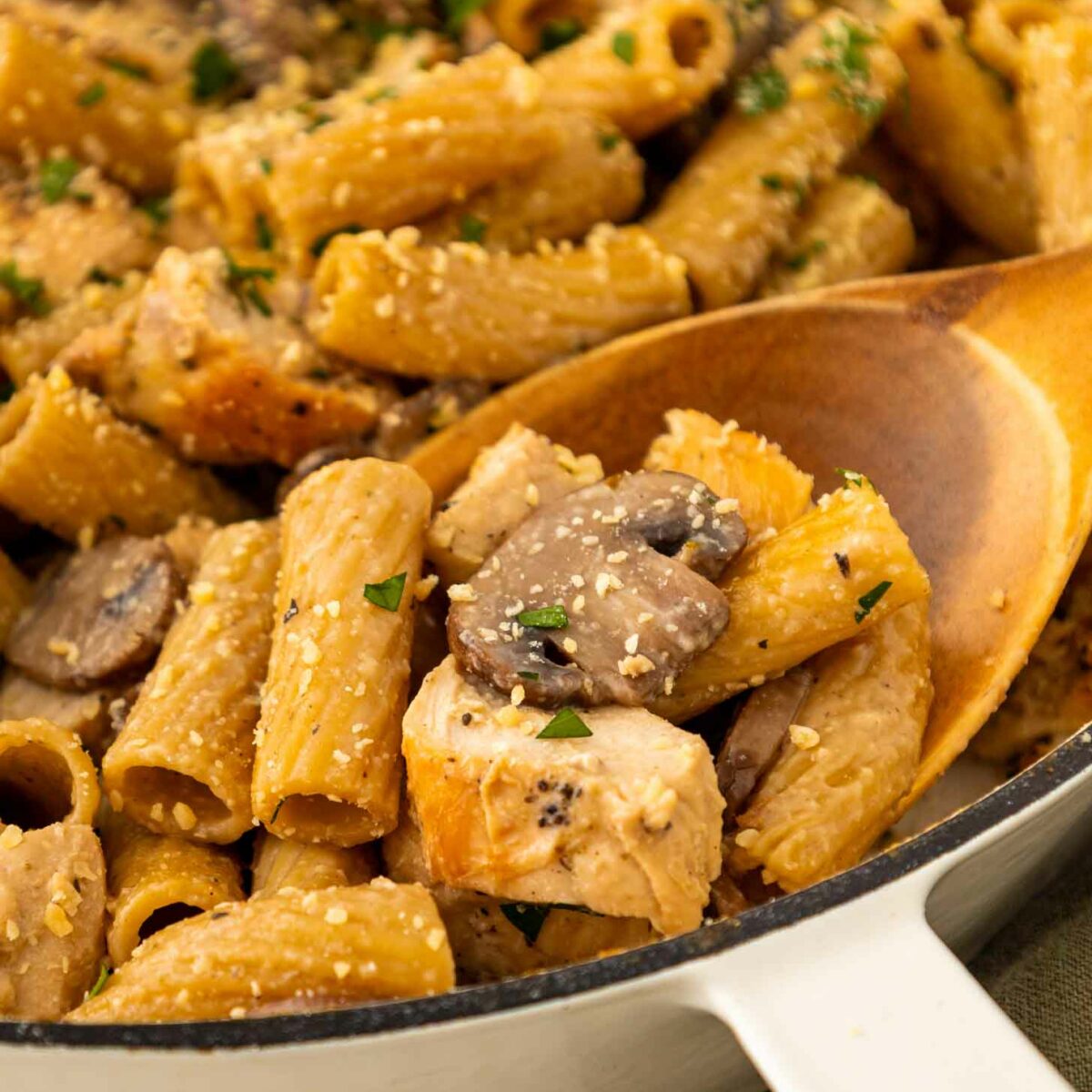 A close-up of creamy rigatoni pasta with chunks of chicken, sliced mushrooms, and a sprinkle of herbs and grated cheese, served in a white skillet with a wooden spoon.