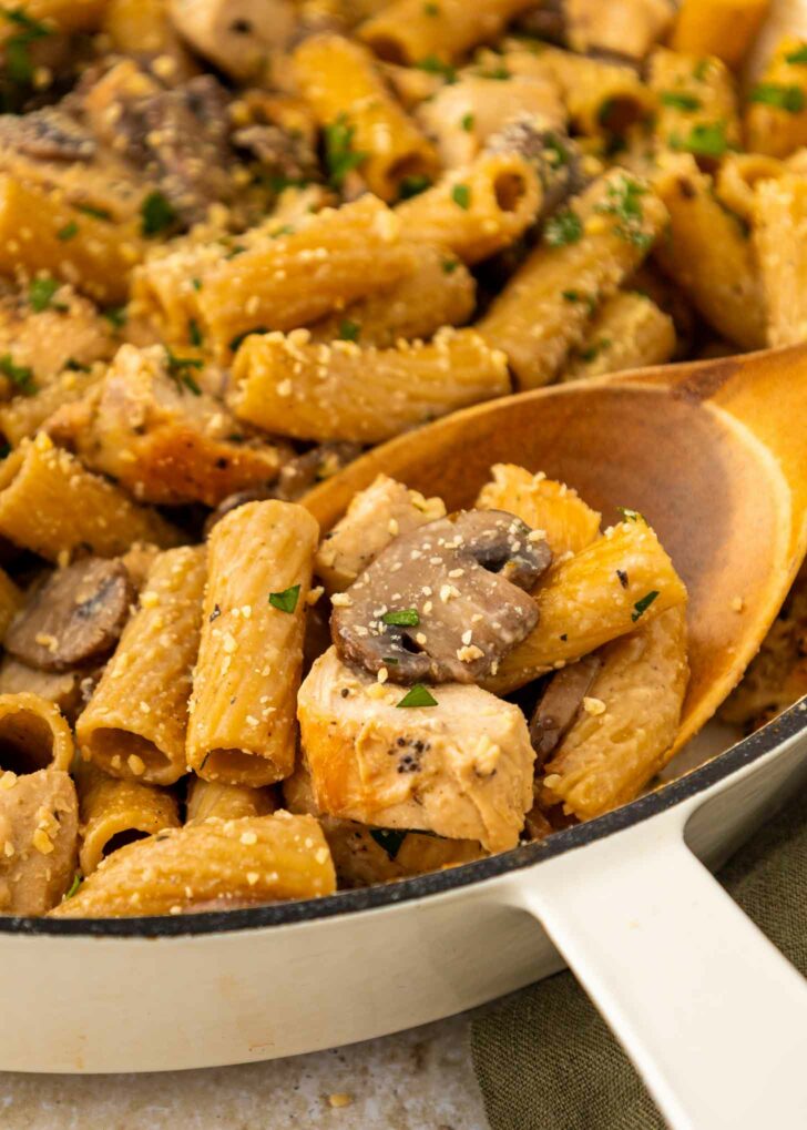A close-up of creamy rigatoni pasta with chunks of chicken, sliced mushrooms, and a sprinkle of herbs and grated cheese, served in a white skillet with a wooden spoon.