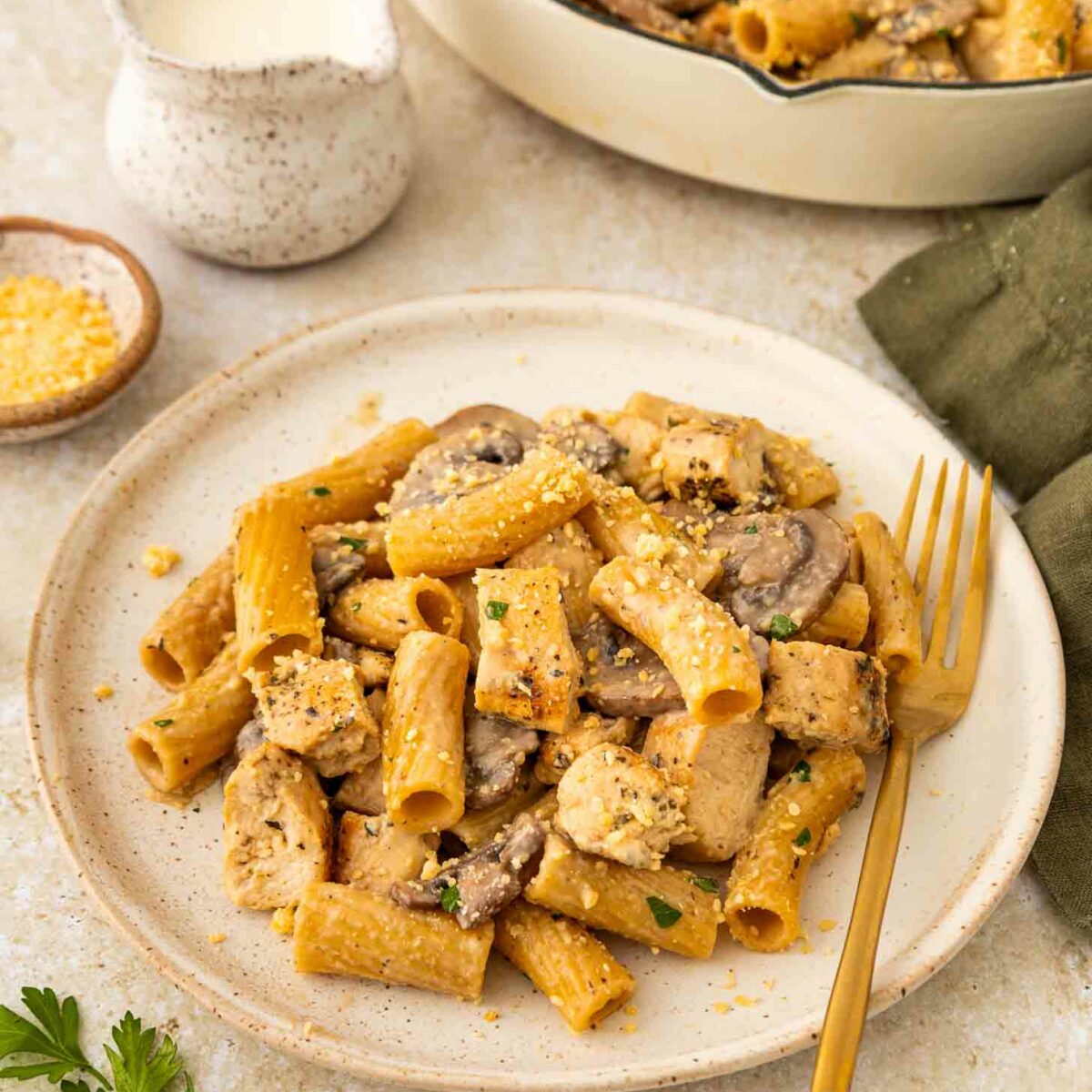 A plate of rigatoni pasta with sliced mushrooms and pieces of chicken in a creamy sauce, garnished with herbs, sits beside a gold fork. A green napkin, grated cheese, and a small pitcher are nearby.