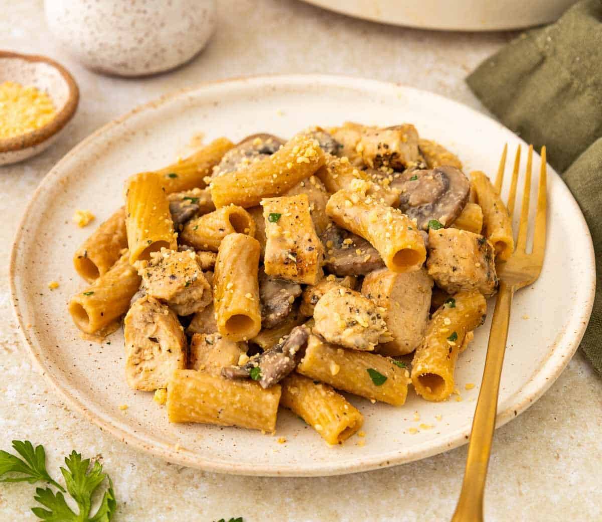 A plate of rigatoni pasta mixed with pieces of chicken and mushrooms, garnished with herbs, sits on a beige surface. A gold fork, a small bowl of grated cheese, and a green napkin are nearby.