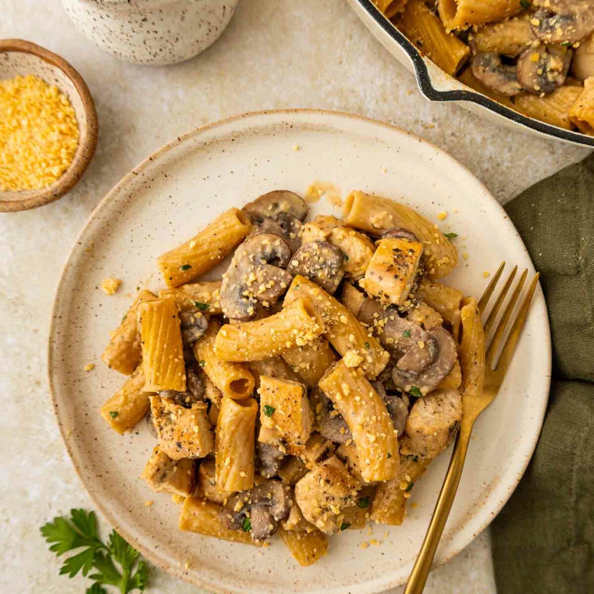 A plate of rigatoni pasta with pieces of chicken, mushrooms, and a creamy sauce, garnished with grated cheese. A gold fork rests on the plate. Nearby are a jug of sauce, a small bowl of cheese, and fresh parsley.
