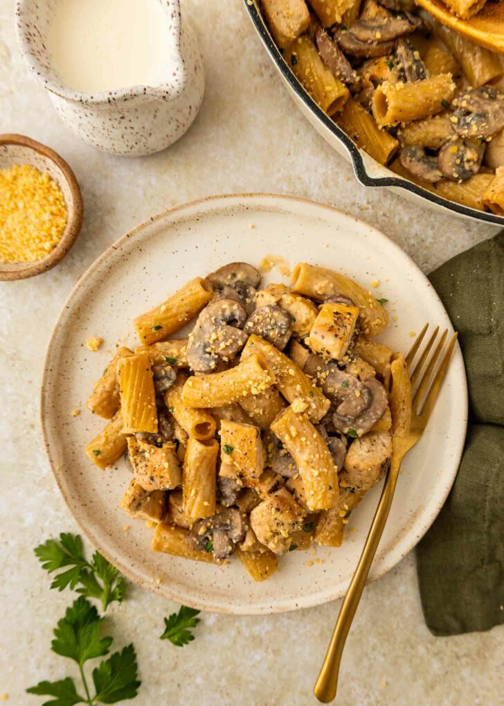 A plate of rigatoni pasta with pieces of chicken, mushrooms, and a creamy sauce, garnished with grated cheese. A gold fork rests on the plate. Nearby are a jug of sauce, a small bowl of cheese, and fresh parsley.