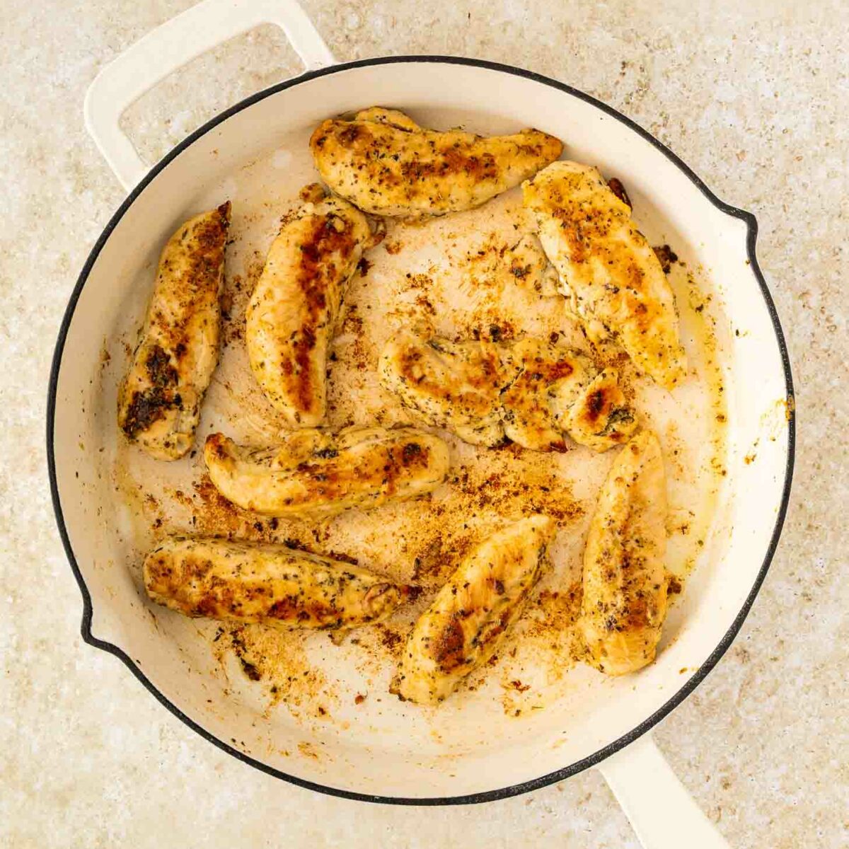 Sautéed chicken tenders seasoned with spices are cooking in a white skillet on a light-colored countertop. The chicken is golden brown with visible seasoning.