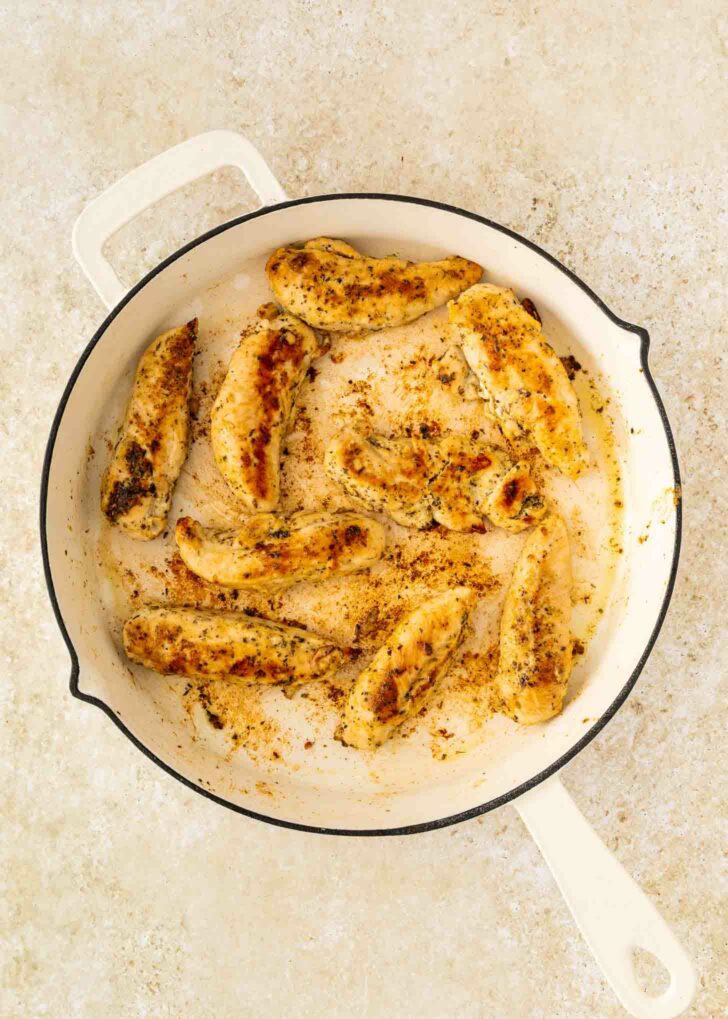 Sautéed chicken tenders seasoned with spices are cooking in a white skillet on a light-colored countertop. The chicken is golden brown with visible seasoning.