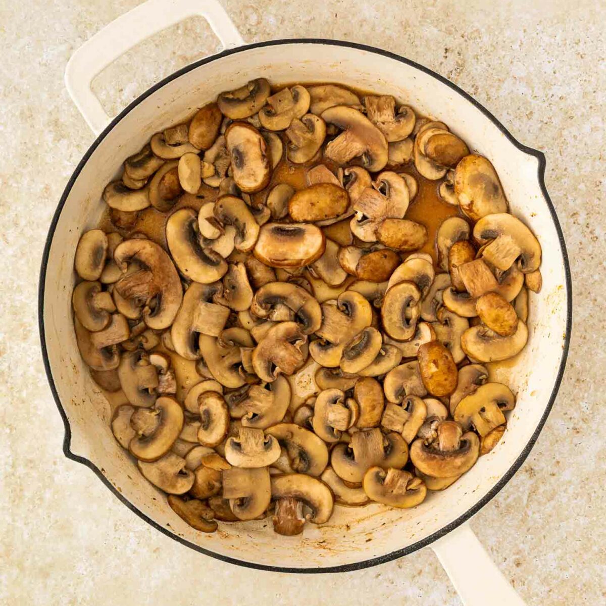 A white skillet filled with sautéed sliced mushrooms on a light, textured countertop surface.