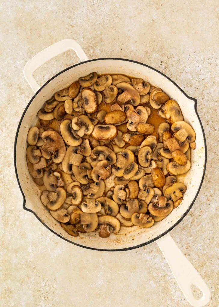 A white skillet filled with sautéed sliced mushrooms on a light, textured countertop surface.