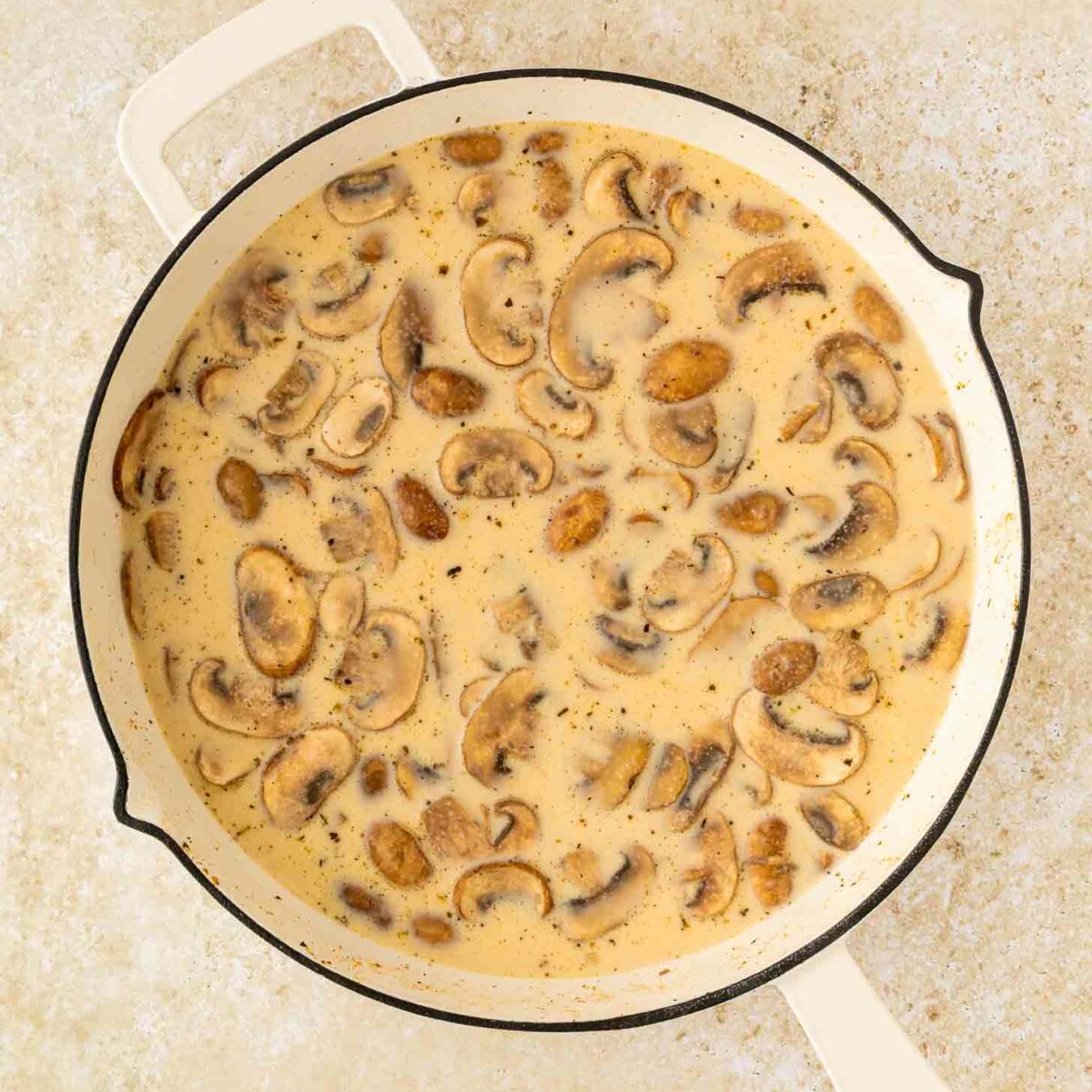 A white skillet filled with creamy mushroom sauce, showing sliced mushrooms in a thick, beige-colored sauce on a light, textured surface.
