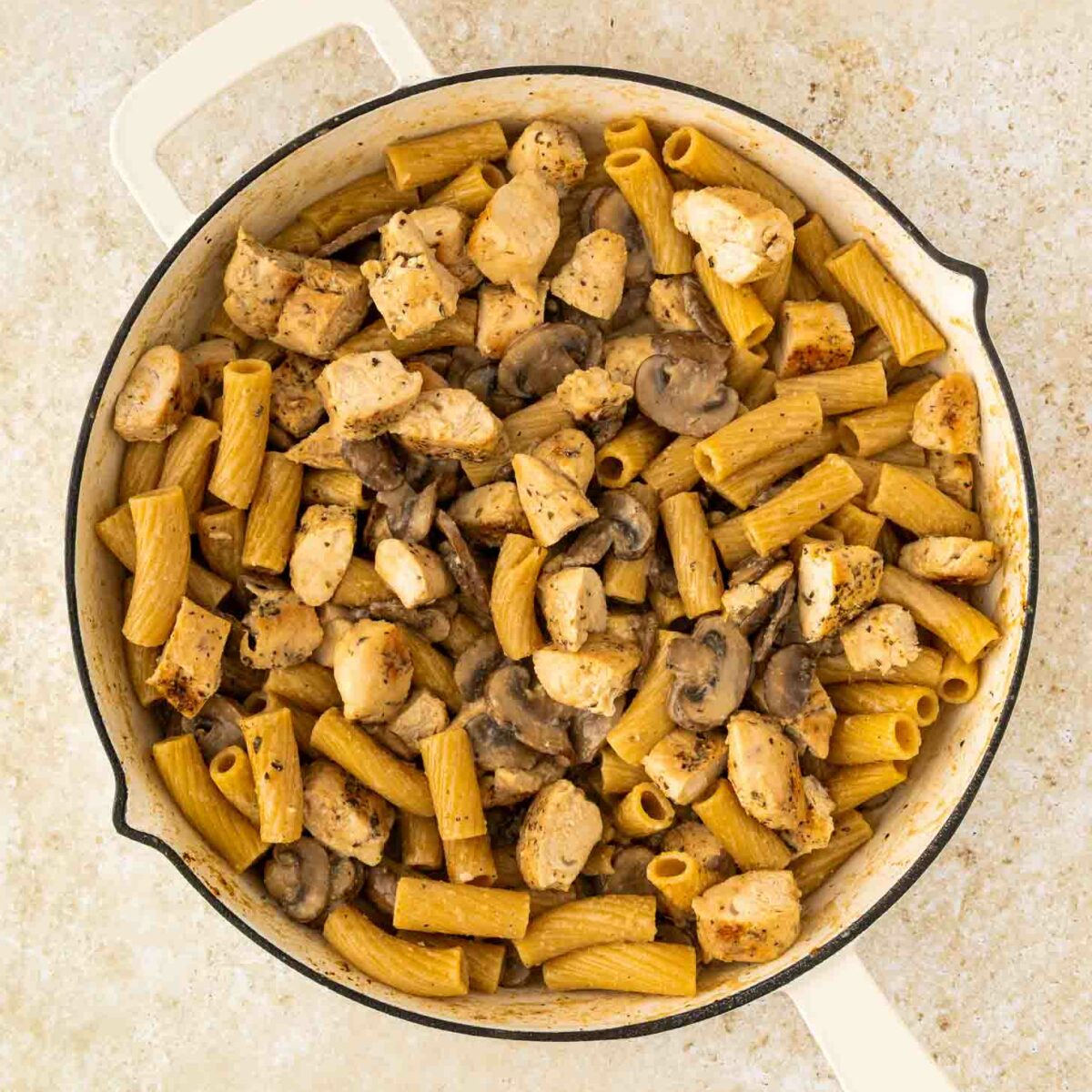 A white pot filled with rigatoni pasta, diced cooked chicken, and sliced mushrooms, all mixed together, sits on a light-colored countertop.