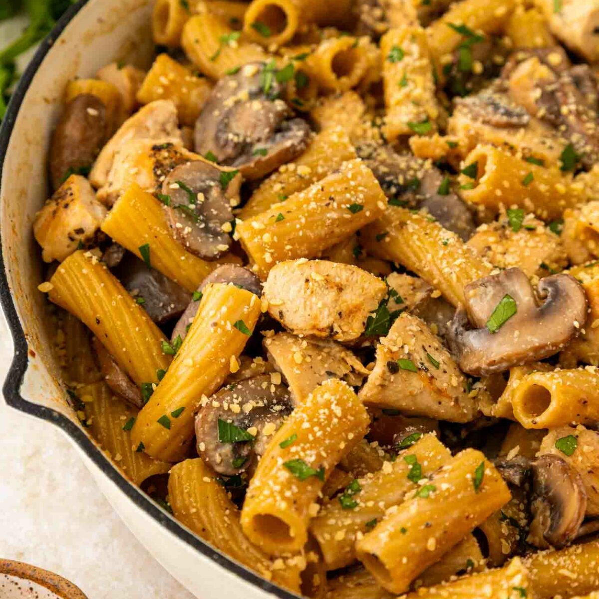 A close-up of a chicken mushroom skillet filled with rigatoni pasta, cooked chicken pieces, mushrooms, fresh parsley, and sprinkled grated parmesan cheese.