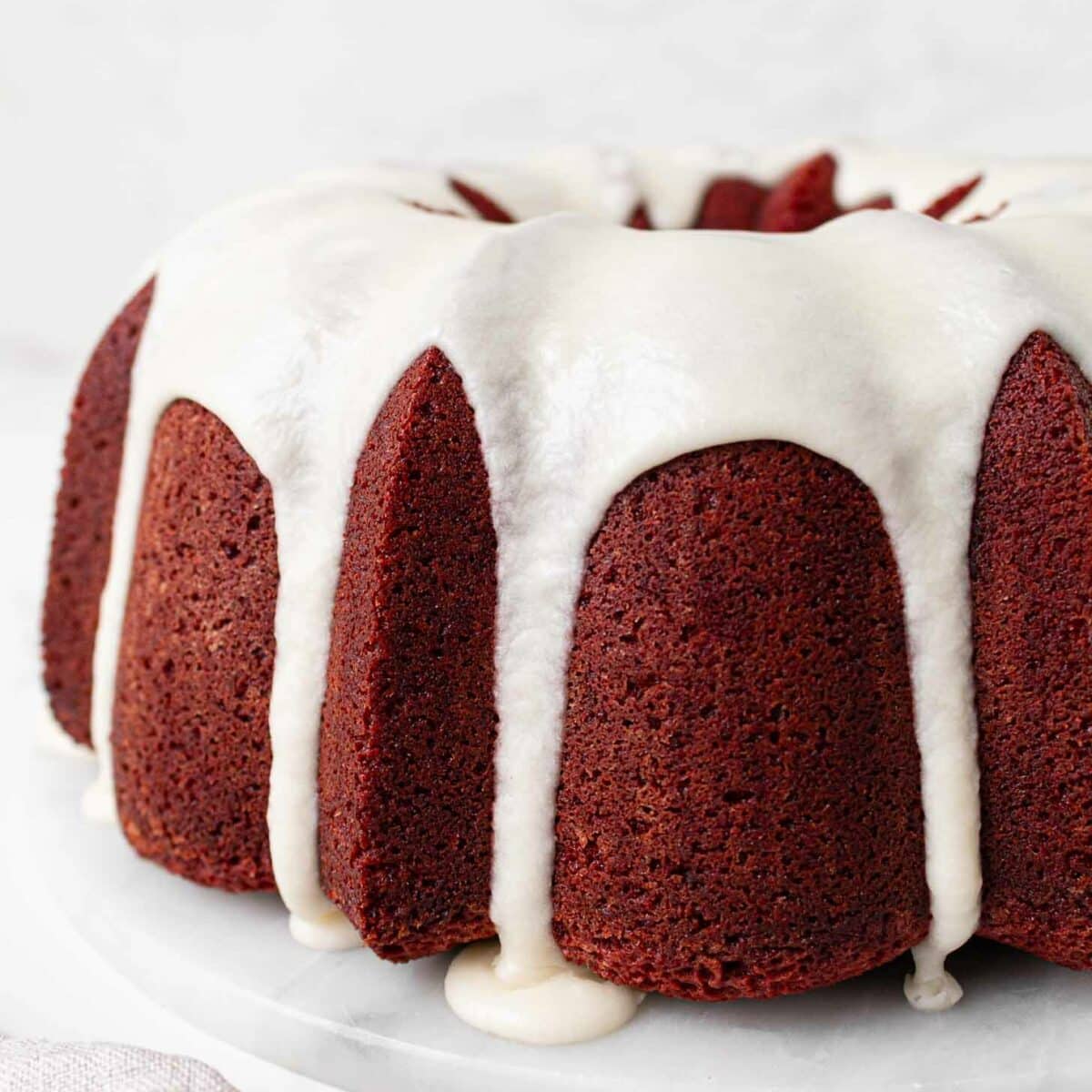 A red velvet pound cake topped with white cream cheese icing, which drips down the sides, is displayed on a white marble plate against a light background.