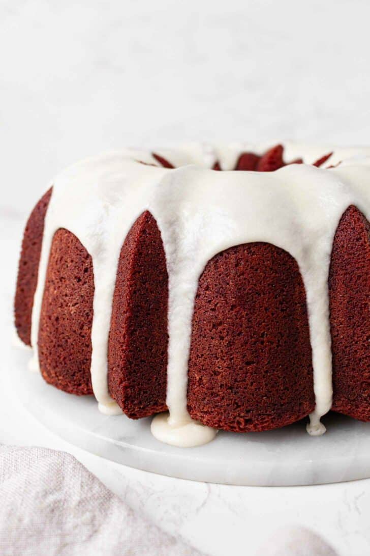 A red velvet pound cake topped with white cream cheese icing, which drips down the sides, is displayed on a white marble plate against a light background.