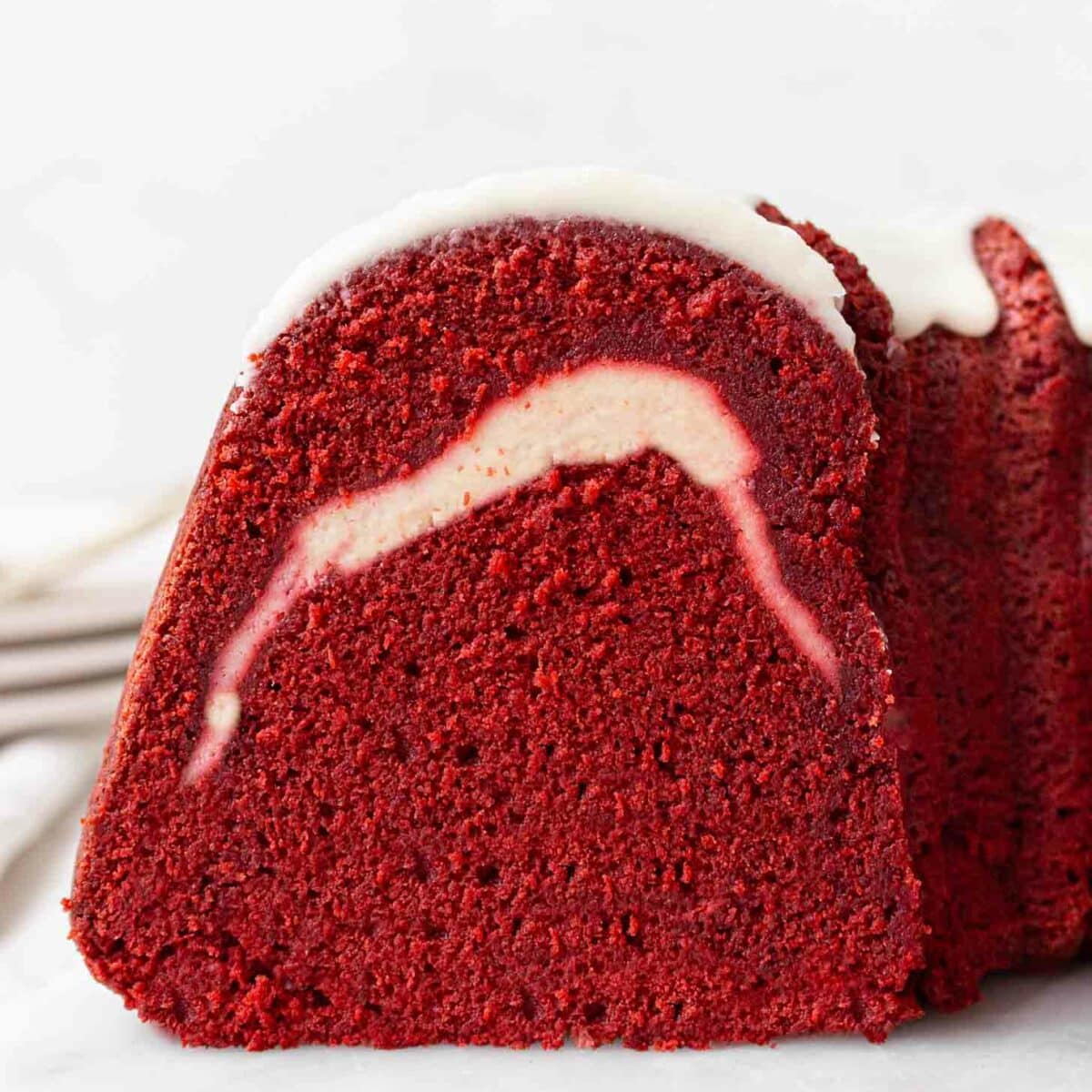 A close-up of a slice of red velvet pound cake with a cream cheese swirl in the center and white icing drizzled on top, set on a white surface.