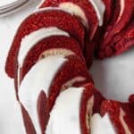 A sliced red velvet pound cake topped with white icing, arranged in a ring shape on a white surface. Plates and a fork are visible in the background.