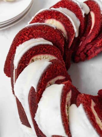 A sliced red velvet pound cake topped with white icing, arranged in a ring shape on a white surface. Plates and a fork are visible in the background.