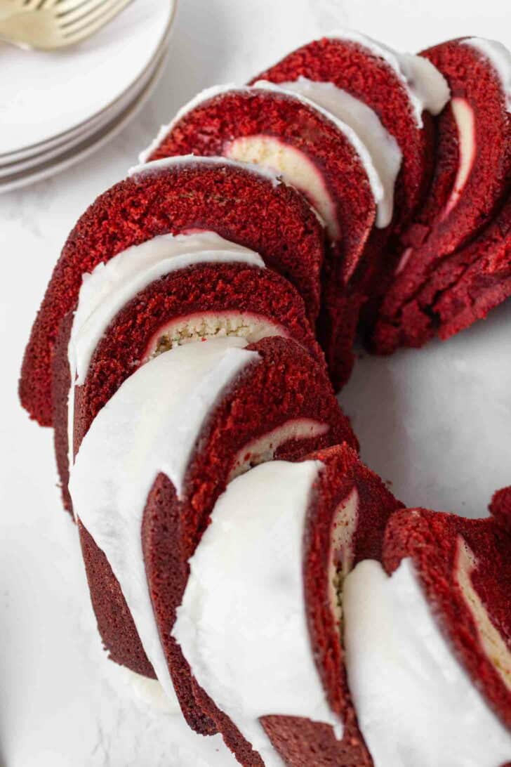 A sliced red velvet pound cake topped with white icing, arranged in a ring shape on a white surface. Plates and a fork are visible in the background.