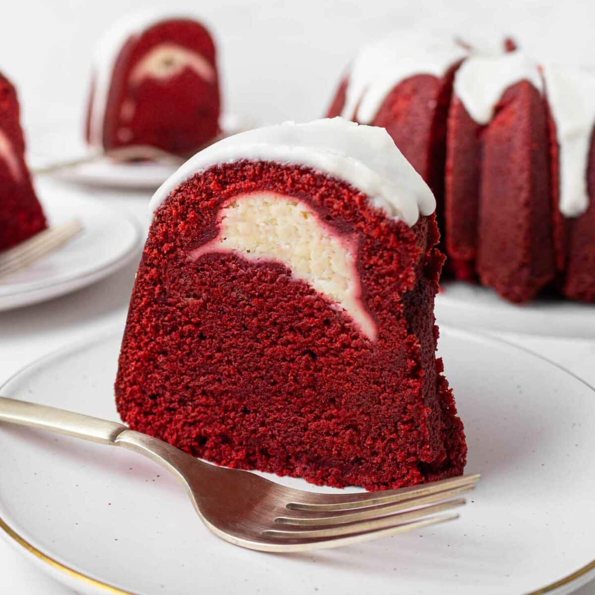 A slice of red velvet Bundt cake with a cream cheese filling and white icing on top, served on a white plate with a fork. The whole cake and another slice are in the background.