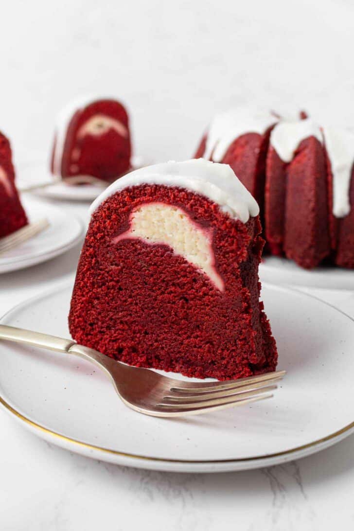 A slice of red velvet Bundt cake with a cream cheese filling and white icing on top, served on a white plate with a fork. The whole cake and another slice are in the background.
